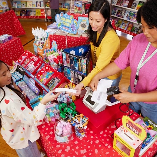 One child is at a cashier table using eWallet. The table is covered with colorful books and stationery. Books cases filled with books are in the background.