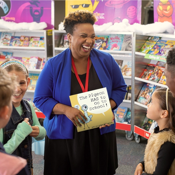 A teacher smiles while showing off a book to their students with Book Fair cases in the background