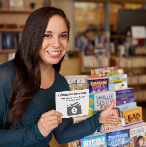 A smiling teacher gives a thumbs up next to a stack of new books from the Book Fair 