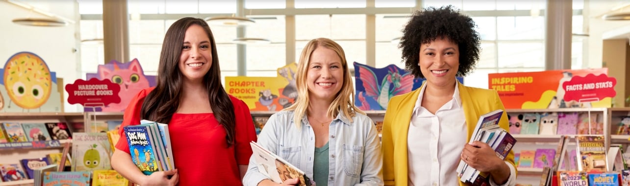 Three Book Fair Hosts smile in front of fully stocked Book Far cases holding books from their Fair