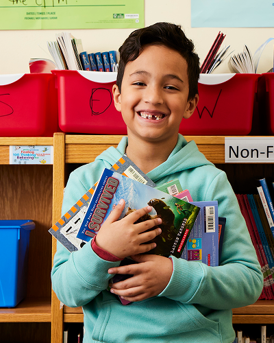 Child in a light blue hoodie holding books in front of a 'Non-Fiction' bookshelf with red letter-labeled bins.