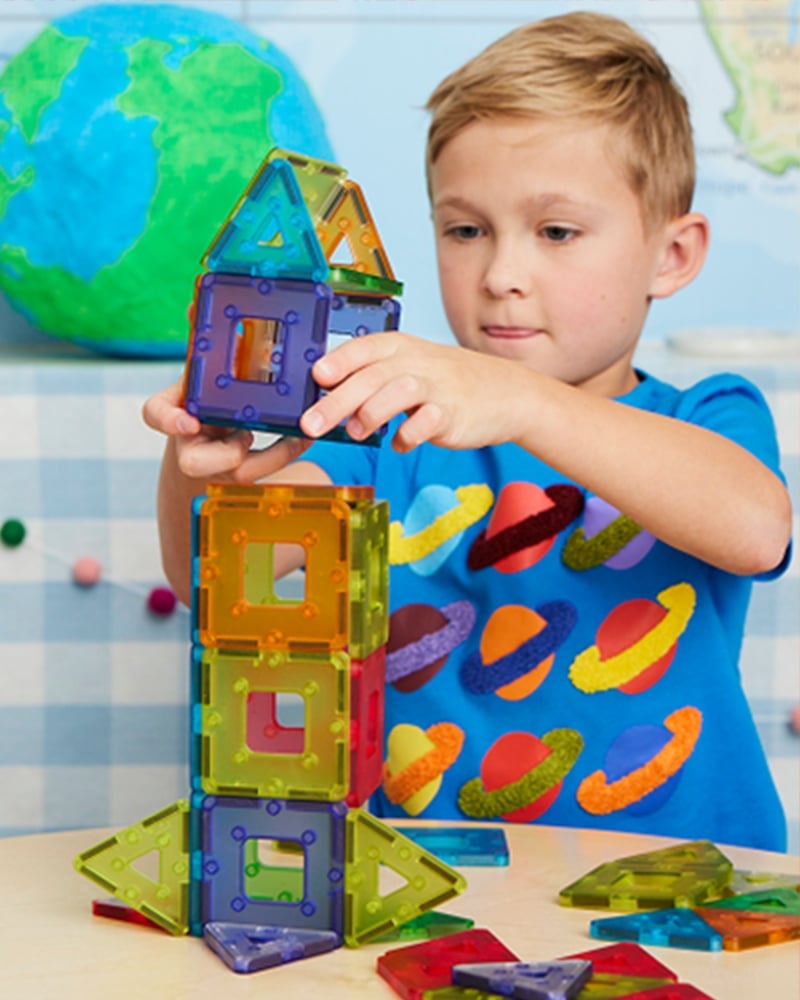 Child in a blue shirt with planet designs building a colorful magnetic tile tower on a table. A globe and partial world map are visible in the background.