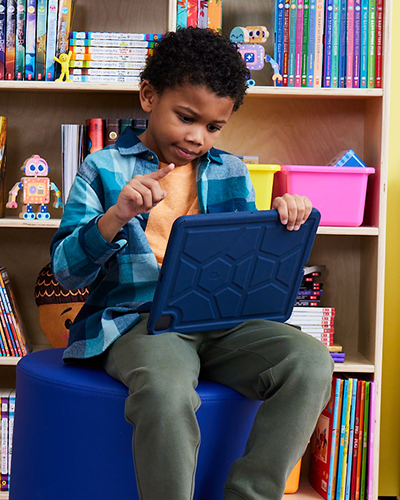 Child sitting on a blue stool using a blue tablet in front of a bookshelf filled with colorful books and toys.