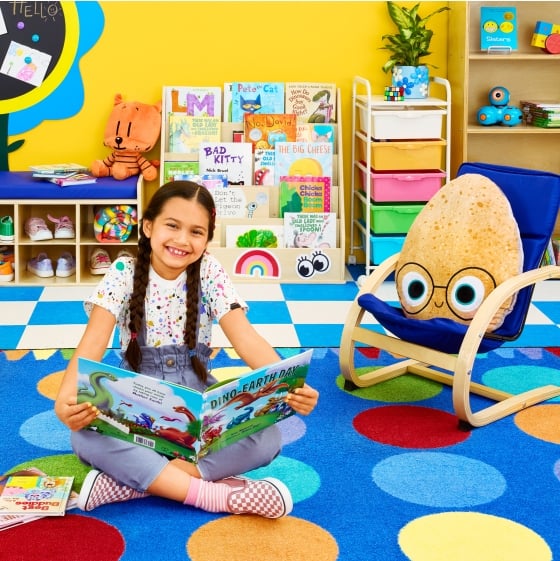 A student smiles while reading a book surrounded by Catalog available books, furniture, rugs, and characters